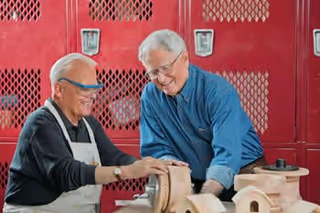 Two elderly men working together on a woodworking project in a workshop with red lockers in the background. One man is wearing safety glasses and an apron, while the other is assisting him.