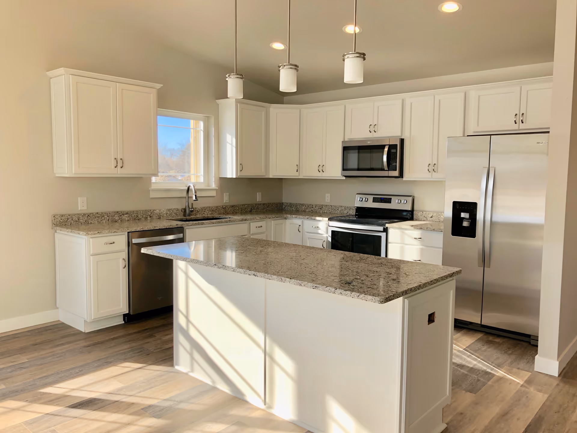 Bright modern kitchen with white cabinets, granite countertops, stainless steel appliances including a refrigerator, microwave, stove, and dishwasher. There is a kitchen island in the center with pendant lights hanging above. The floor is wood-style and there is a window letting in natural light.