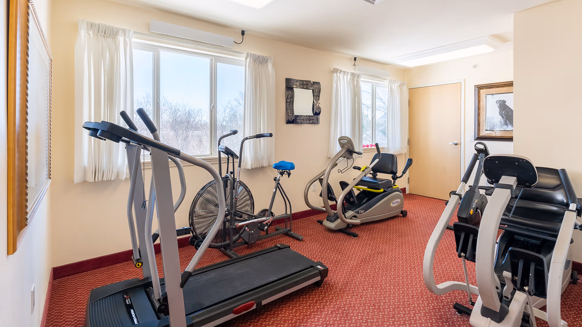 A small fitness room with exercise equipment including a treadmill, stationary bike, recumbent bike, and a multi-function weight machine. The room has red carpet, beige walls, two windows with white curtains, a framed mirror, and a framed picture on the wall.