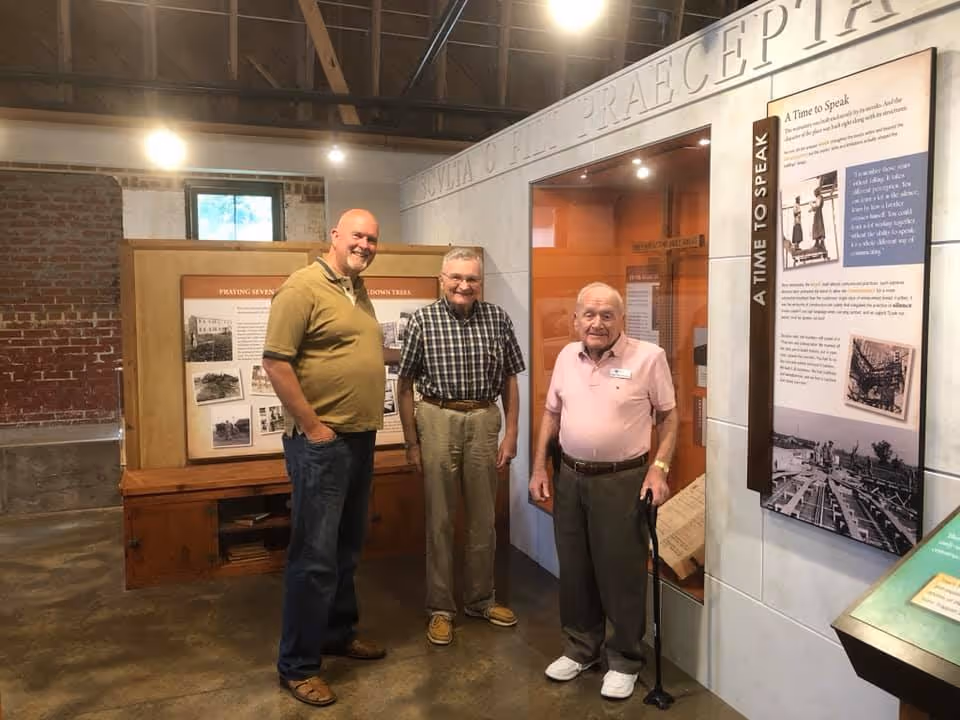 Three men standing inside a museum or exhibit area with informational displays on the walls. One man is wearing a yellow shirt and jeans, the second man is wearing a checkered shirt and khaki pants, and the third man is wearing a pink polo shirt, gray pants, and holding a cane. The background includes brick walls, a window, and exhibit panels with text and images.