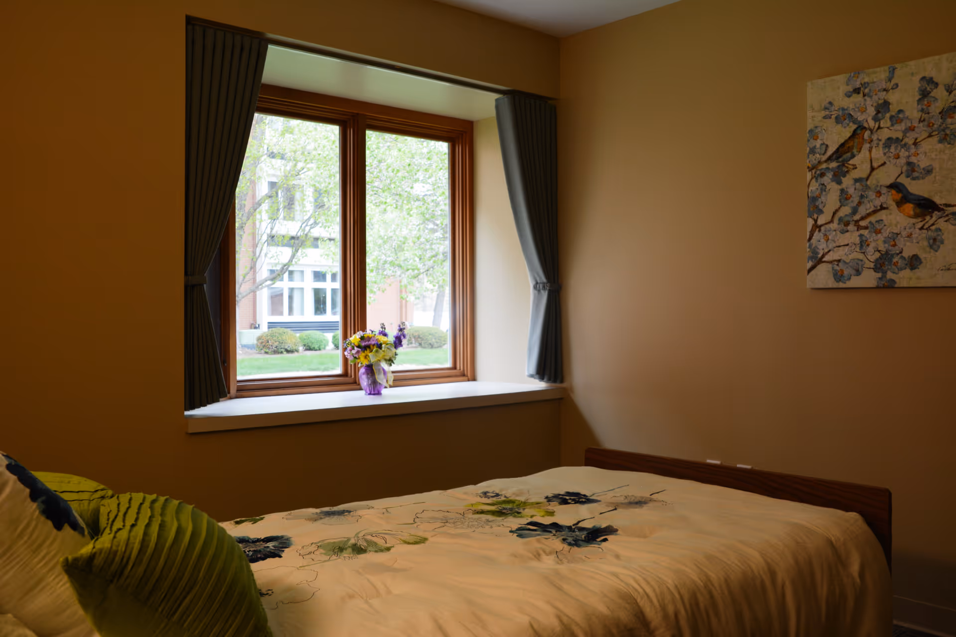 A cozy bedroom with a single bed covered in a floral-patterned bedspread. There are green and blue pillows on the bed. A window with gray curtains tied back lets in natural light and shows a view of trees and a building outside. A painting of birds on a branch hangs on the beige wall.