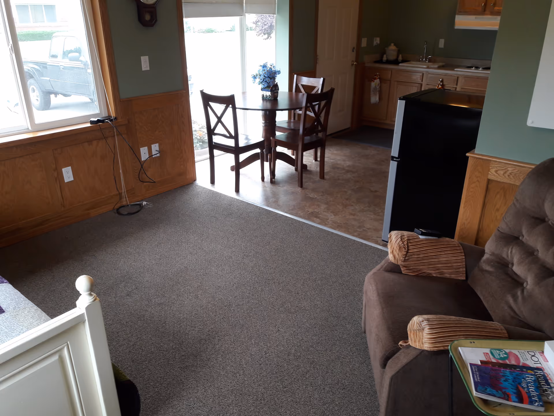 Interior view of a retirement home showing a small dining area with a round wooden table and three chairs near a sliding glass door. Adjacent to the dining area is a kitchenette with wooden cabinets, a sink, and a small black refrigerator. In the foreground, there is a brown cushioned armchair with striped armrest covers and a side table holding magazines. A window with a view of a parked vehicle is on the left side, and part of a white bed frame is visible in the bottom left corner.