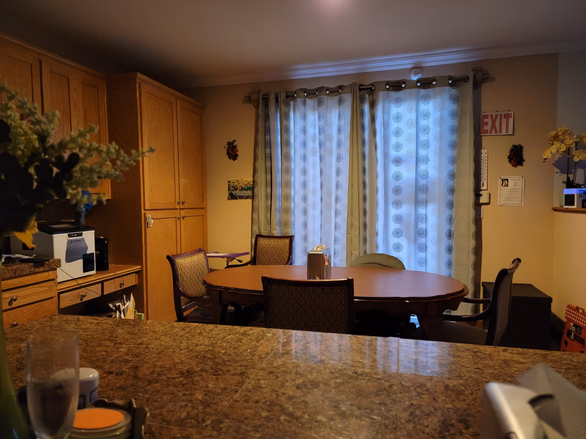 Interior view of a dining area with a wooden table surrounded by chairs. Behind the table are large windows covered with patterned curtains. To the left, there are wooden cabinets and a countertop with a coffee machine and other items. An exit sign is visible on the wall to the right.