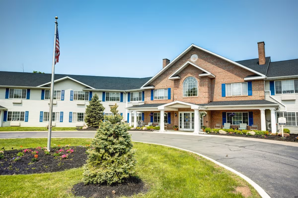 Exterior view of Evergreen Place, a two-story senior living facility with a combination of brick and white siding. The building features blue shutters on the windows, a covered entrance with white columns, and a circular driveway. In front of the building, there is a flagpole with an American flag, landscaped greenery, and small trees under a clear blue sky.