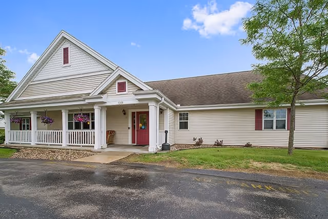 Exterior view of a single-story building with beige siding and a gabled roof. The building has a covered front porch with white columns and hanging flower baskets. There is a red front door and a small tree on the right side of the building. The area in front includes a paved driveway and a grassy lawn.