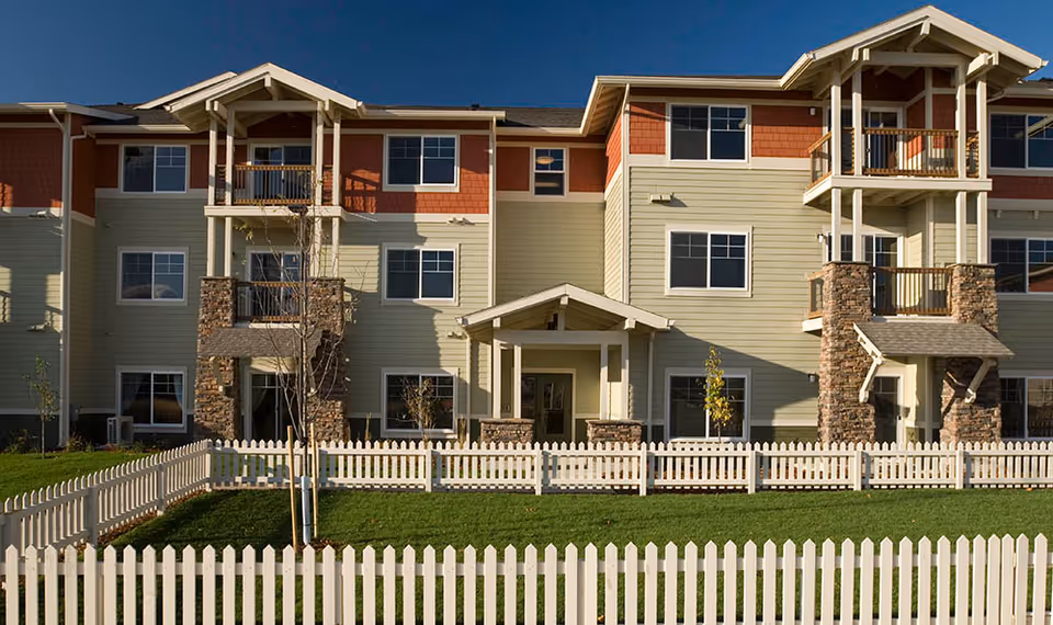 A multi-story senior living facility with a front porch and stone accents, surrounded by a white picket fence.
