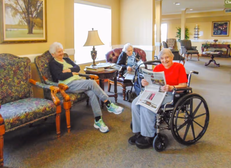 Three elderly women in a well-lit common area of a senior living facility. One woman in a red shirt is sitting in a wheelchair reading a newspaper and smiling. Another woman is seated on a floral-patterned armchair with her arms crossed, and the third woman is sitting on a leather chair with a walker nearby. The room has beige walls, carpeted floor, a table with a lamp, and framed artwork on the walls.