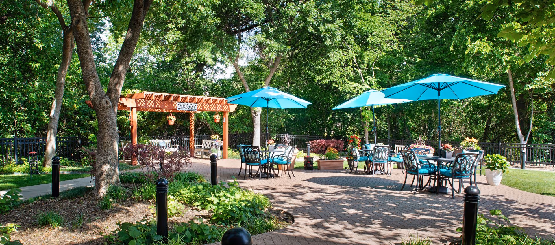 Sunlit outdoor patio with teal umbrellas over tables and chairs, a wooden pergola, and surrounding trees and landscaping.