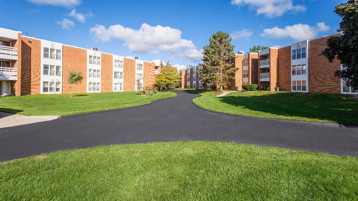 Exterior view of a senior living facility with two three-story brick buildings facing each other across a paved driveway. The area is surrounded by well-maintained green lawns, a few trees, and a partly cloudy blue sky.