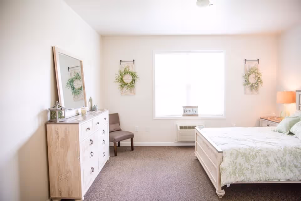 A bright and airy bedroom with a large window letting in natural light. The room features a white wooden bed with green and white bedding, a matching dresser with a large mirror, a brown chair, two decorative wreaths on the wall, and a bedside table with a lamp.
