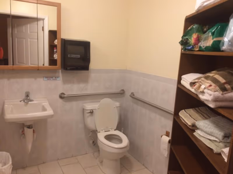 A small bathroom with a white toilet with the lid open, a white wall-mounted sink, and a wooden shelving unit filled with folded towels and supplies. The walls are tiled halfway up with light-colored tiles and painted beige above. There are metal grab bars installed on the walls near the toilet for accessibility. A mirror cabinet and a black paper towel dispenser are mounted above the sink.