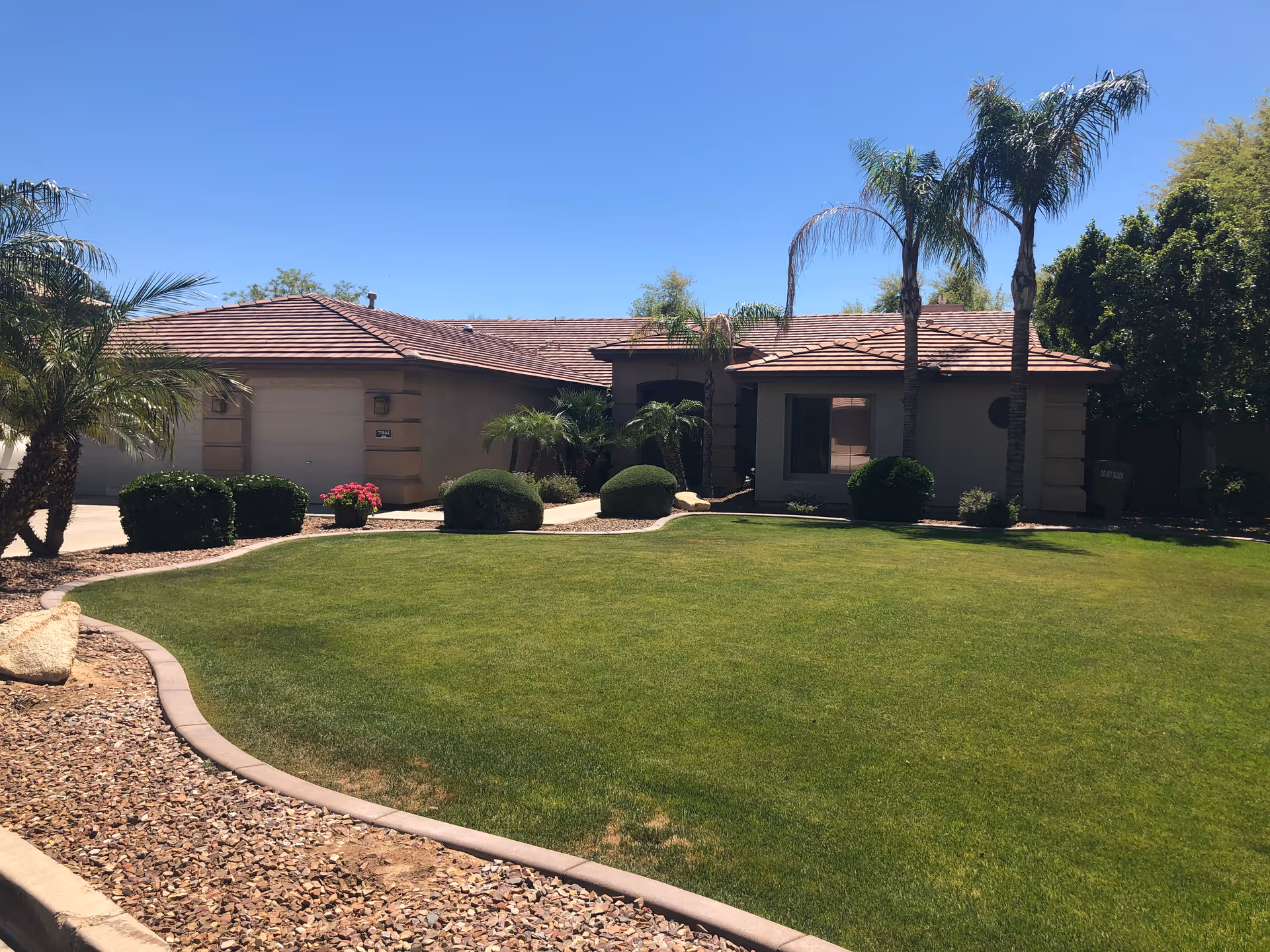 Single-story stucco house front with a manicured lawn, palm trees, and a driveway under a clear blue sky.