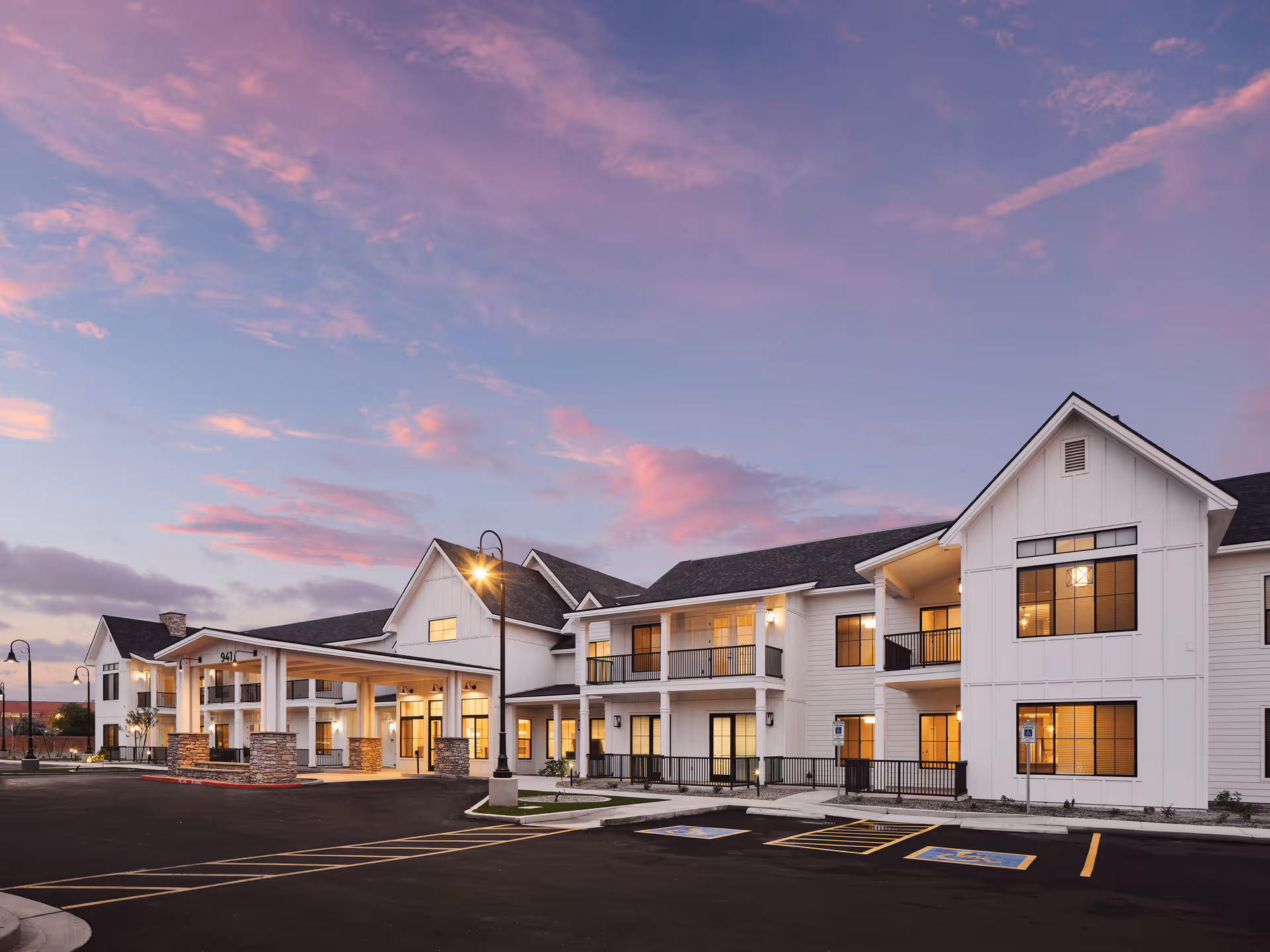 Exterior view of a modern senior living facility building at dusk with warm interior lights visible through large windows, a covered entrance with stone pillars, and a parking lot with handicap spaces in front.
