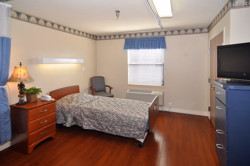 A simple bedroom in a senior living facility with a single bed covered in a floral patterned quilt, a wooden nightstand with a lamp and a small plant, a gray armchair, a window with blinds and a blue valance, a wall-mounted air conditioning unit, and a blue dresser with a flat-screen TV on top. The room has wood flooring and beige walls with a decorative border near the ceiling.