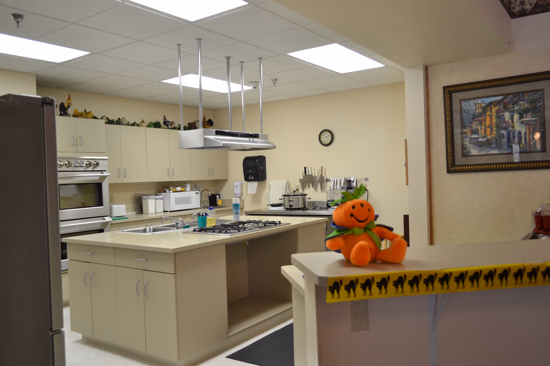 Interior view of a kitchen in an assisted living facility featuring beige cabinets, a stove with a range hood, a microwave, a double sink, and various kitchen utensils. A small orange pumpkin plush toy with a smiling face and green scarf sits on the counter in the foreground. A framed painting hangs on the wall to the right.