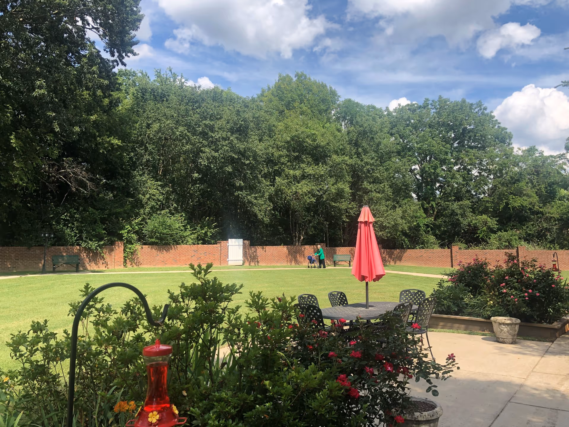 Outdoor garden area with green grass, trees, and a brick wall in the background. There is a patio with a table, chairs, and a closed red umbrella. Flowering bushes and potted plants are visible around the patio. A person is seen walking with a walker near the brick wall.
