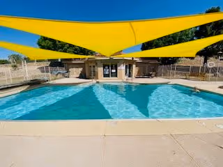 Outdoor swimming pool with clear blue water under large yellow shade sails. There is a small building or pool house at the far end of the pool, surrounded by a fence and trees in the background.