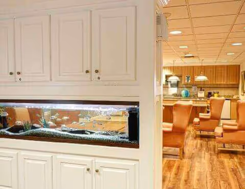 Interior view of a senior living facility showing a built-in aquarium embedded in white cabinetry on the left, with a seating area featuring brown leather armchairs and wooden flooring visible in the background.