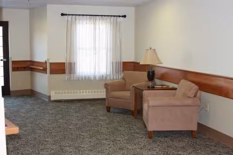 A senior living facility living room with two beige armchairs, a side table, lamp, and window.