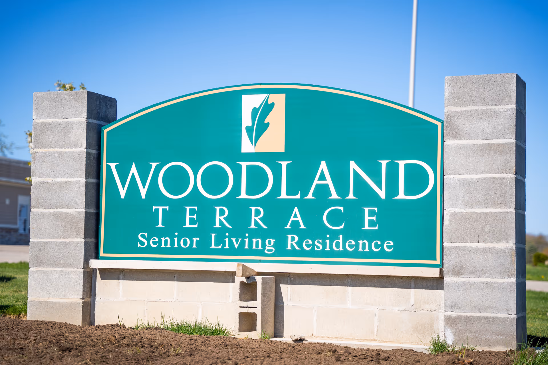 Green and beige sign for Woodland Terrace Senior Living Residence mounted on a concrete block base with a clear blue sky in the background.