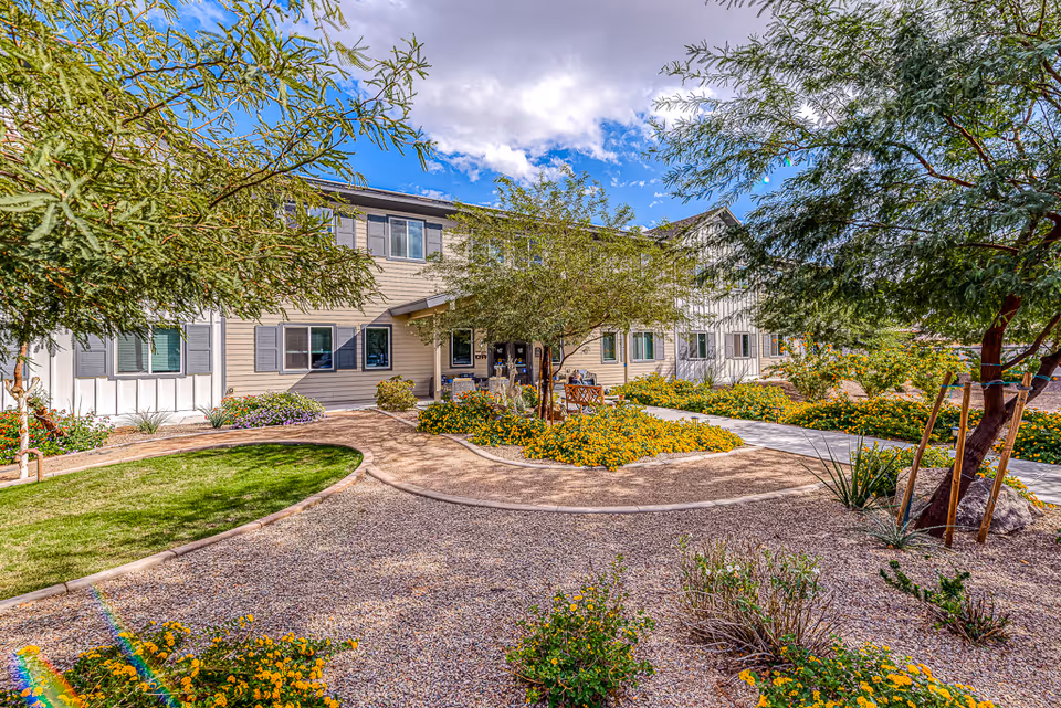 A bright and sunny outdoor garden area at Paradise Valley Senior Living featuring a gravel pathway, green grass patches, yellow flowering plants, and several trees. The two-story building with beige siding and multiple windows is visible in the background under a partly cloudy blue sky.