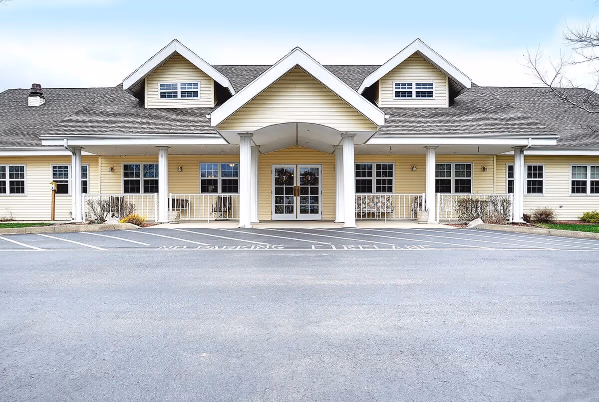 Front exterior view of a single-story building with beige siding, white trim, and a covered entrance supported by four white columns. The building has multiple windows and a gray shingled roof with three dormer windows. The parking area in front has marked no parking fire lane zones.