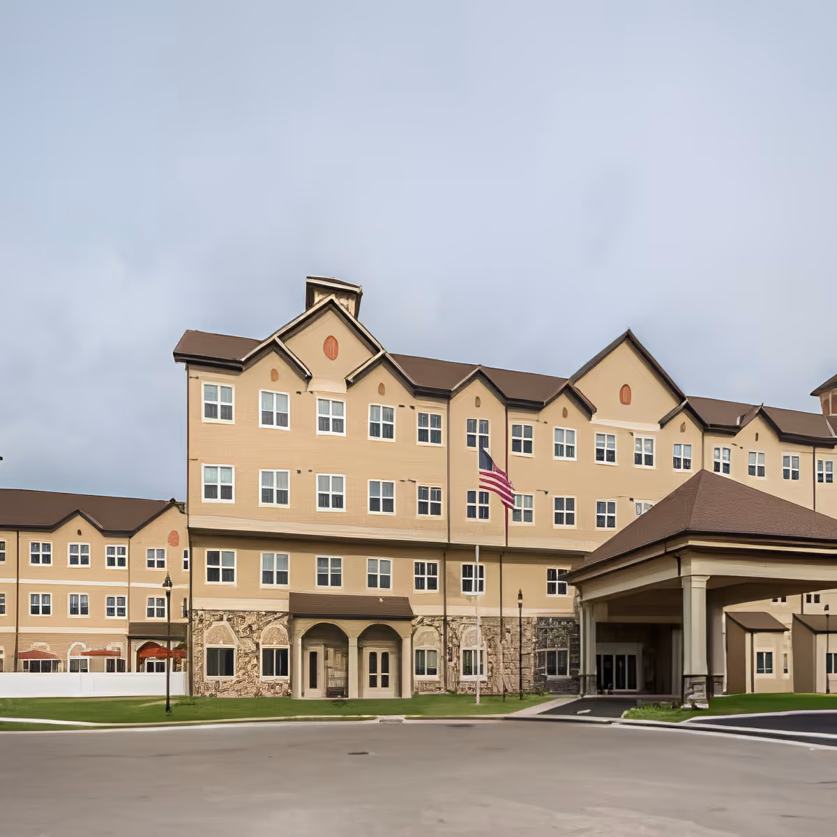 A multi-story beige senior living building with a covered entrance and an American flag flying in front under a cloudy sky.