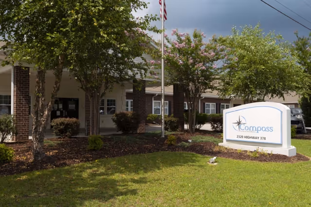 Exterior view of Compass Post Acute Rehabilitation facility showing the front entrance with brick columns, trees, and a sign displaying the facility name and address 2320 Highway 378.