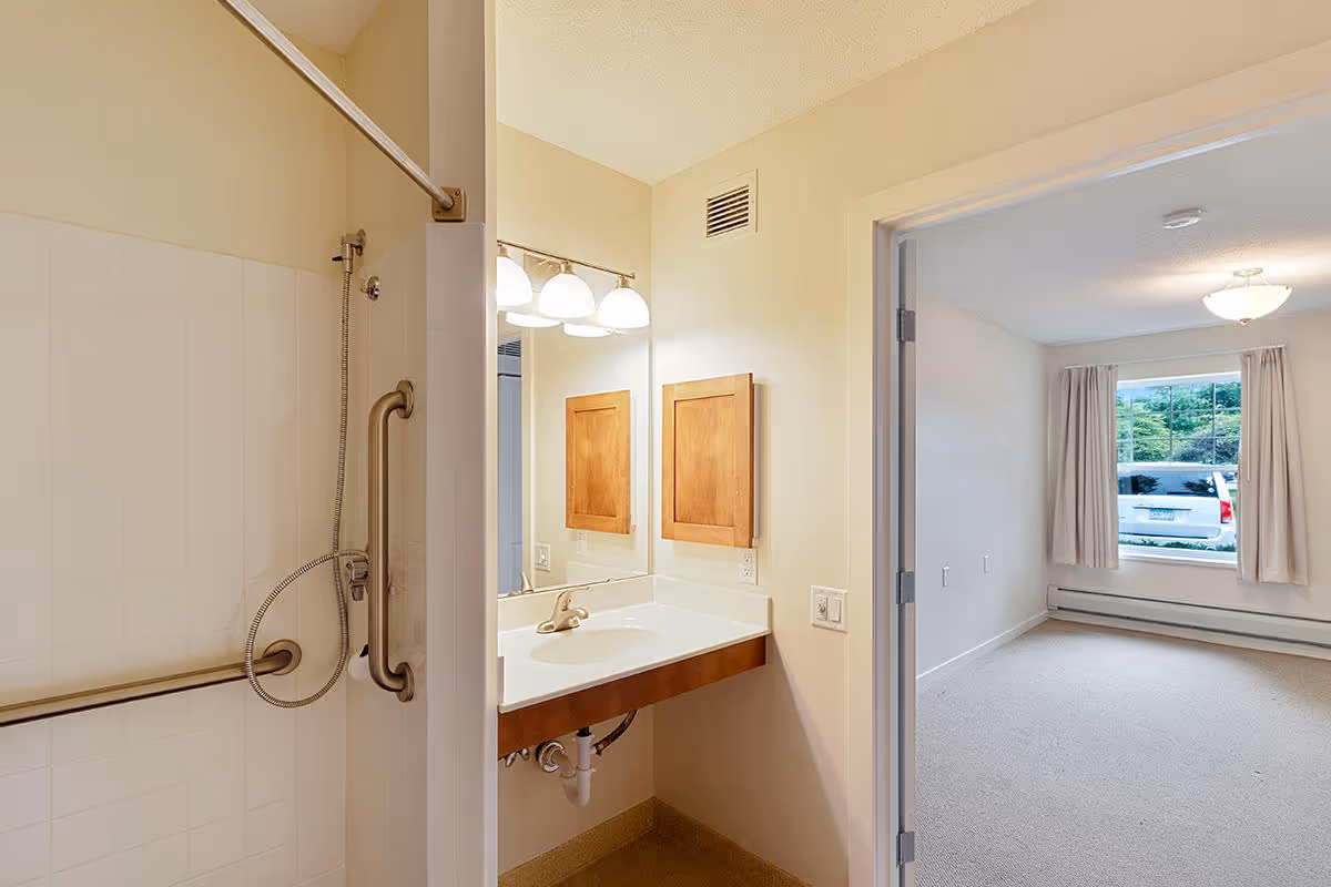 View of a senior housing unit bathroom with a walk-in shower equipped with grab bars and a handheld showerhead. The bathroom has a sink with a large mirror and wooden cabinet above it. Adjacent to the bathroom is a carpeted bedroom with a window showing a parked car outside.