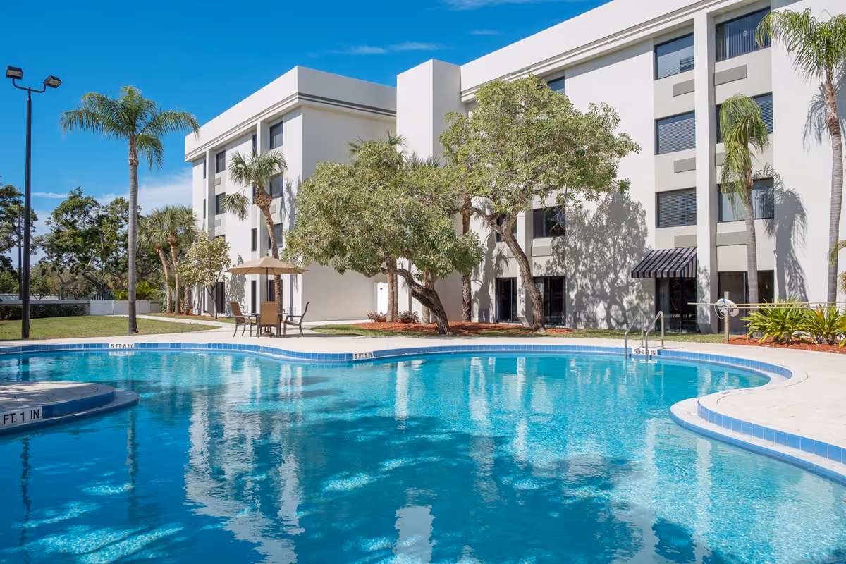 Curved outdoor swimming pool with seating and an umbrella in front of a multi-story white building flanked by palm trees.