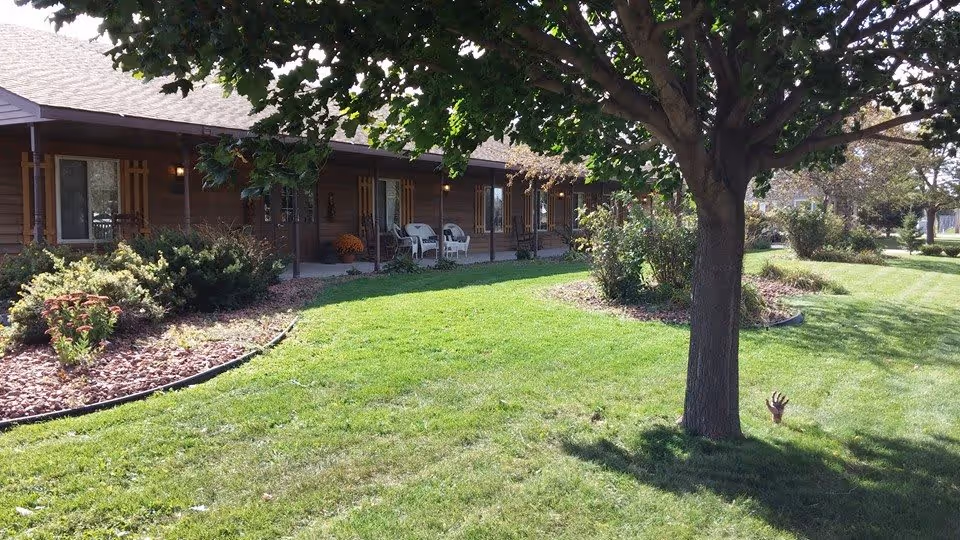 A single-story building with a covered porch featuring white chairs, surrounded by a well-maintained lawn and landscaped garden beds with shrubs and flowers. A large tree stands in the foreground casting shade over the grass.