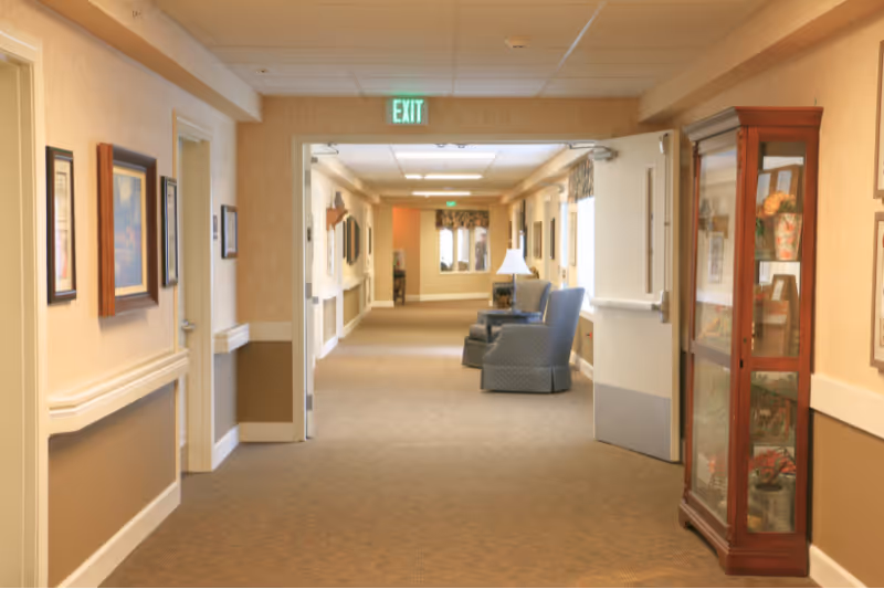 A well-lit hallway in a senior living facility with beige walls and carpeted floor. The hallway features framed artwork on the walls, a glass display cabinet with decorative items on the right, and two upholstered armchairs with a small table and lamp further down the corridor. An exit sign is visible above an open doorway.
