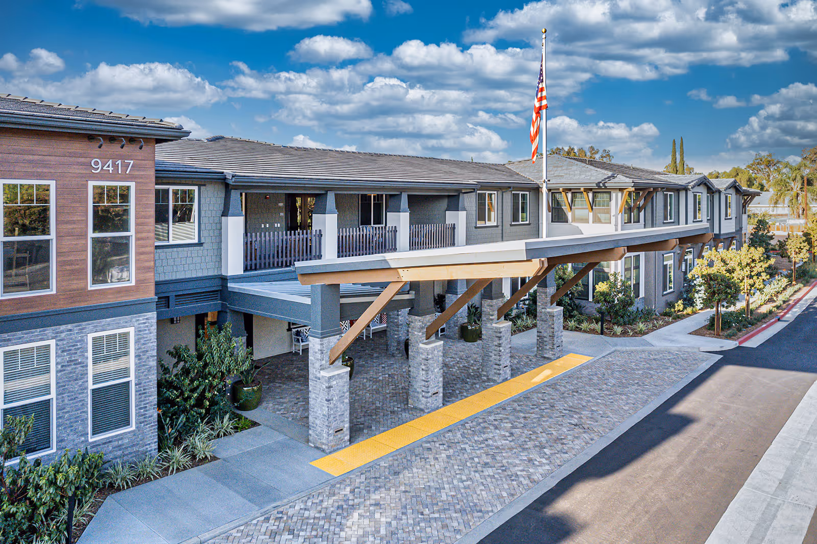 Exterior view of a modern senior living facility building with a covered entrance supported by stone pillars and wooden beams, an American flag on a flagpole, landscaped greenery, and a clear blue sky with scattered clouds.