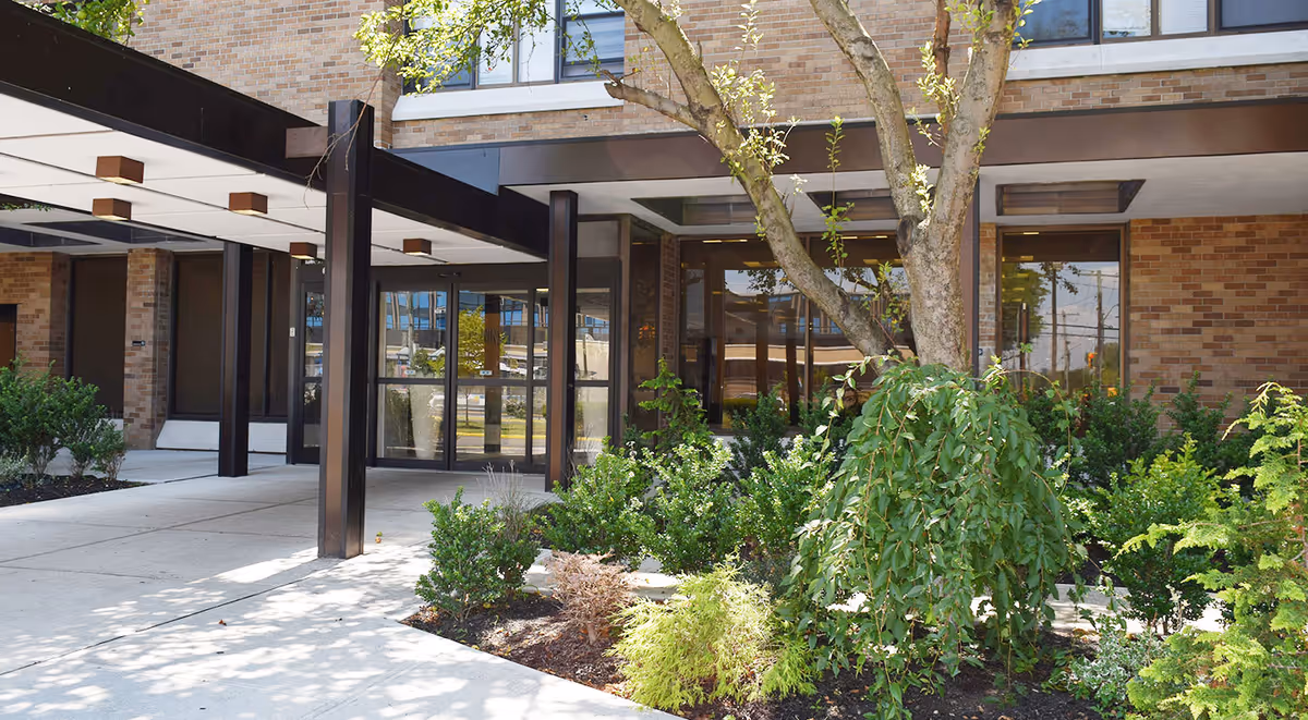 Entrance of a nursing and rehabilitation facility with a covered walkway supported by dark brown beams. There are large glass doors and windows, a brick exterior, and landscaped greenery including bushes and a tree in front of the building.