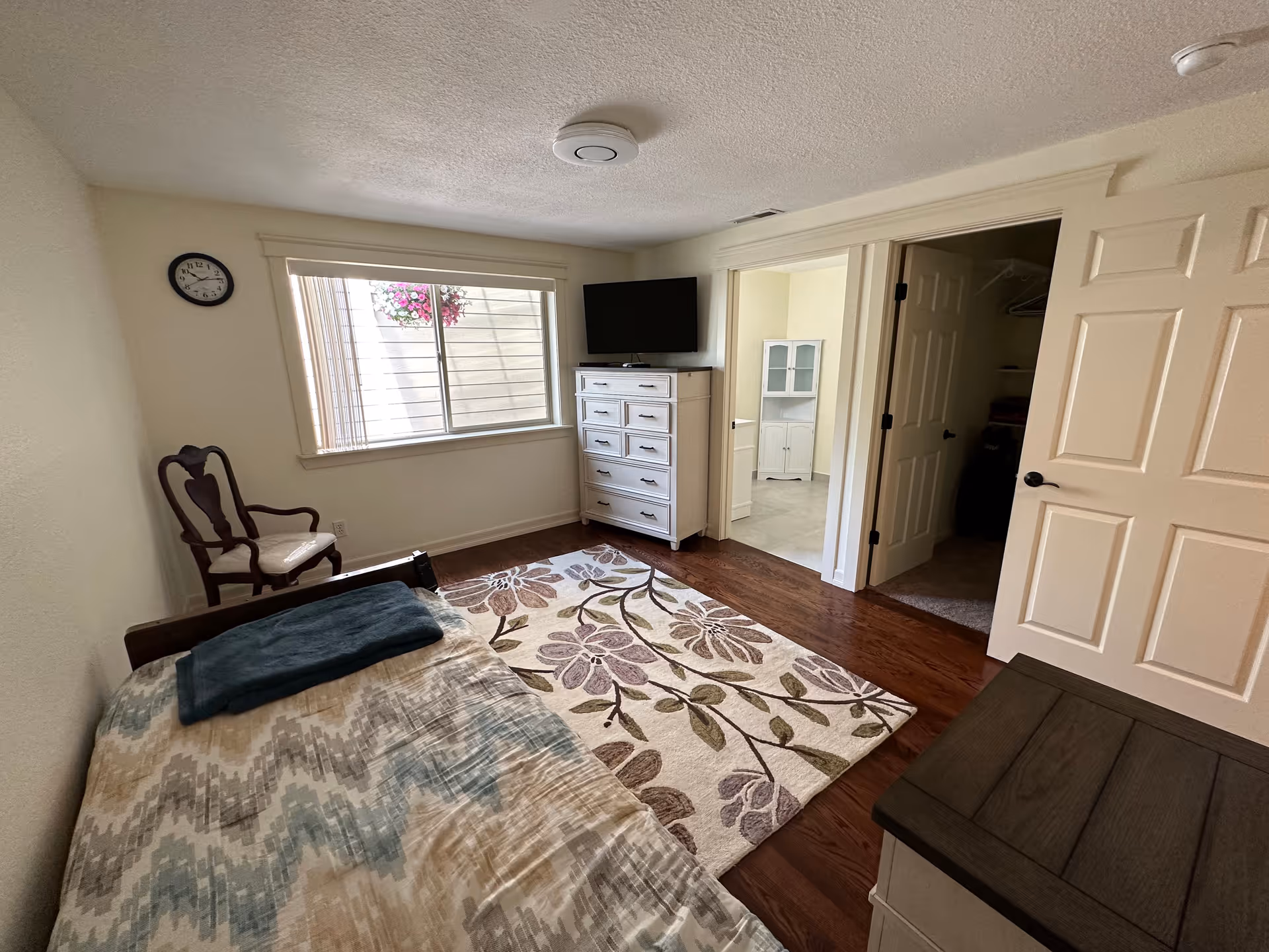 A bedroom with a single bed covered with a patterned blanket and a folded dark blue towel on top. There is a wooden chair with armrests near a window with vertical blinds and a hanging flower basket outside. A white chest of drawers with a flat-screen TV on top is positioned against the wall. The room has wooden flooring with a floral area rug. Two open doors lead to an adjacent room and a closet.
