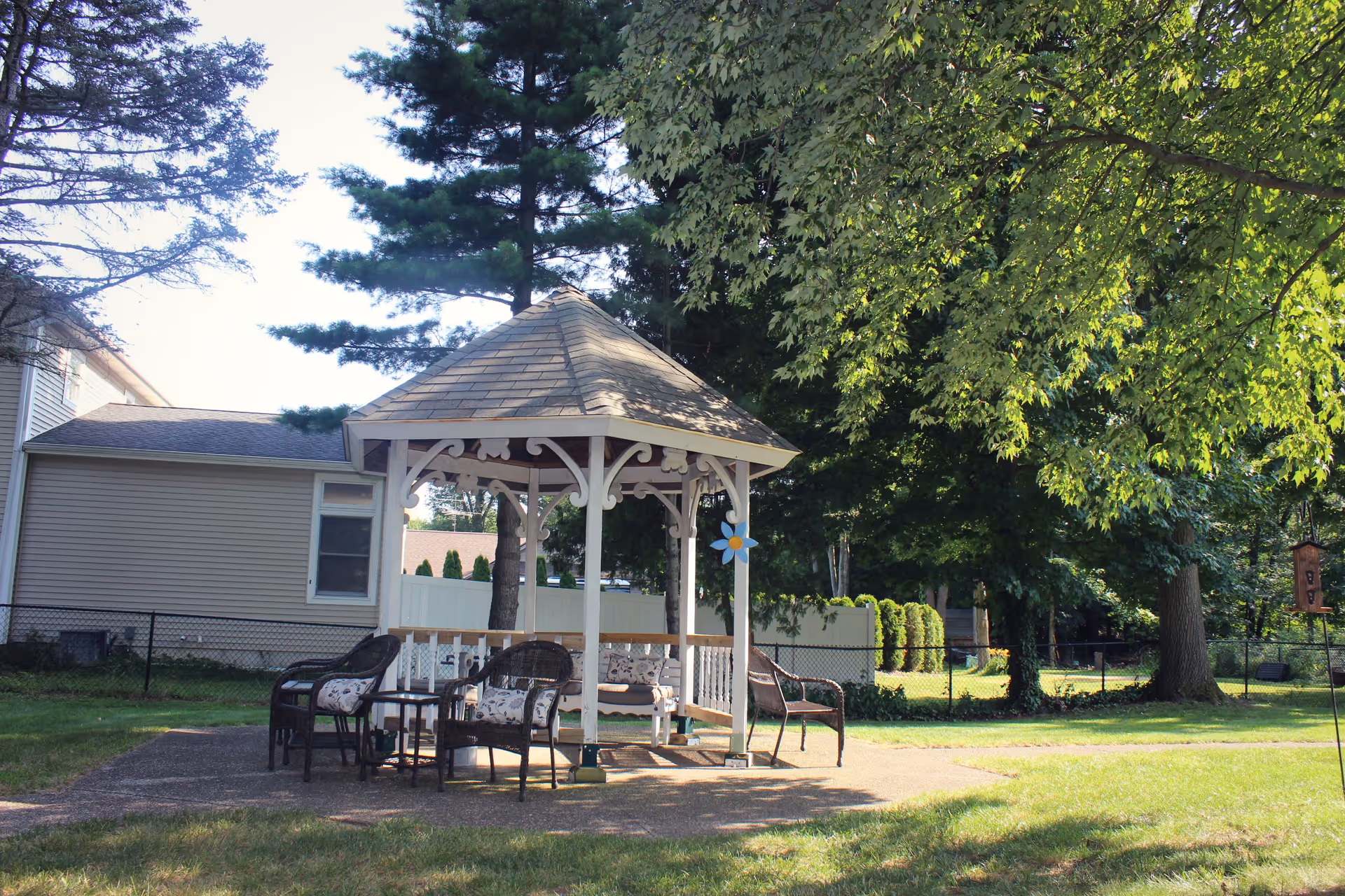 A white wooden gazebo with a shingled roof situated in a grassy outdoor area. The gazebo is furnished with cushioned wicker chairs and a small table. Surrounding the gazebo are trees providing shade, a house with beige siding, and a black chain-link fence enclosing the area.