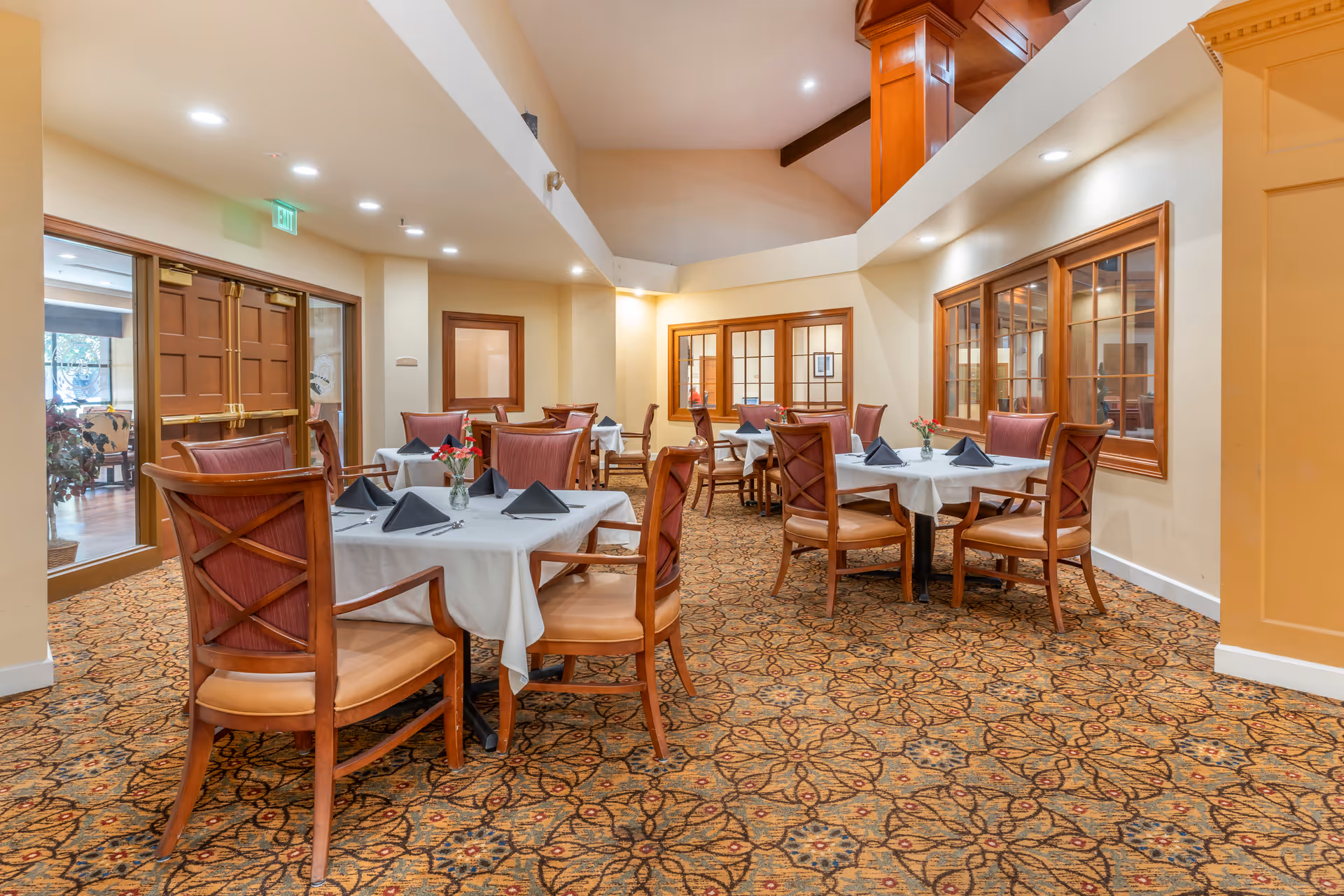 Interior view of a dining room in a senior living facility with several tables covered with white tablecloths, each set with black folded napkins and silverware. The room has patterned carpet flooring, wooden chairs with cushioned seats, and warm lighting. There are large wooden-framed windows and double doors visible in the background.