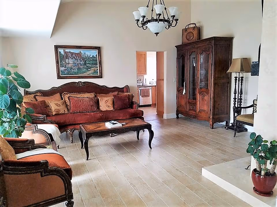 A cozy living room with vintage-style furniture including a maroon sofa with multiple cushions, two armchairs, a wooden coffee table, and a large wooden cabinet. The room has light-colored tiled flooring, a chandelier hanging from the ceiling, a floor lamp, and a framed painting on the wall. There are also two potted plants, one on the left and one on the right near a small raised platform. A doorway leads to a kitchen area in the background.