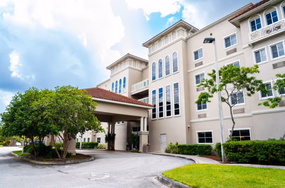 Front exterior of a multi-story senior living building with a covered entrance, landscaped trees, and a driveway.