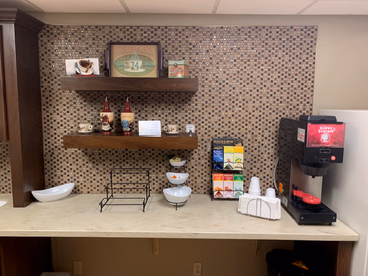 A coffee station with a coffee machine on the right, a display rack with various Bigelow tea bags, disposable cups, napkins, and three white bowls on a beige countertop. Above the counter are two wooden shelves mounted on a mosaic tile wall, holding decorative coffee-themed items including cups, bottles, and signs.