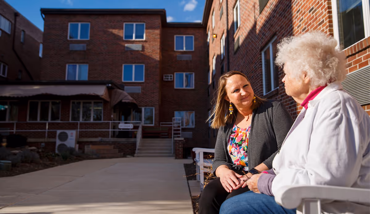 Two women sitting and talking on a bench outside a brick building under a clear blue sky. One woman is elderly with white hair wearing a white jacket, and the other is younger with brown hair wearing a floral top and dark cardigan.