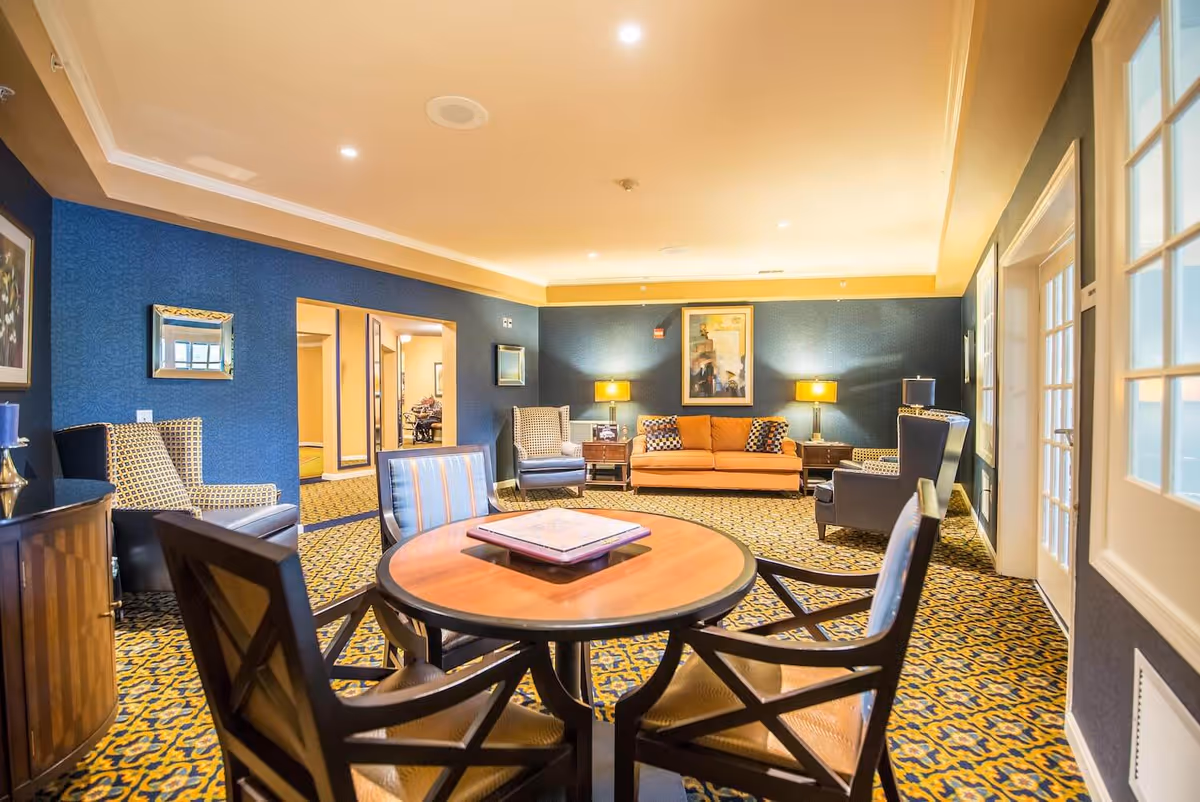 A well-lit living room with patterned carpet, a round wooden table with four chairs in the foreground, and a seating area with an orange sofa, two armchairs, and two side tables with lamps against a blue wall. There is a framed abstract painting above the sofa and a doorway leading to another room.