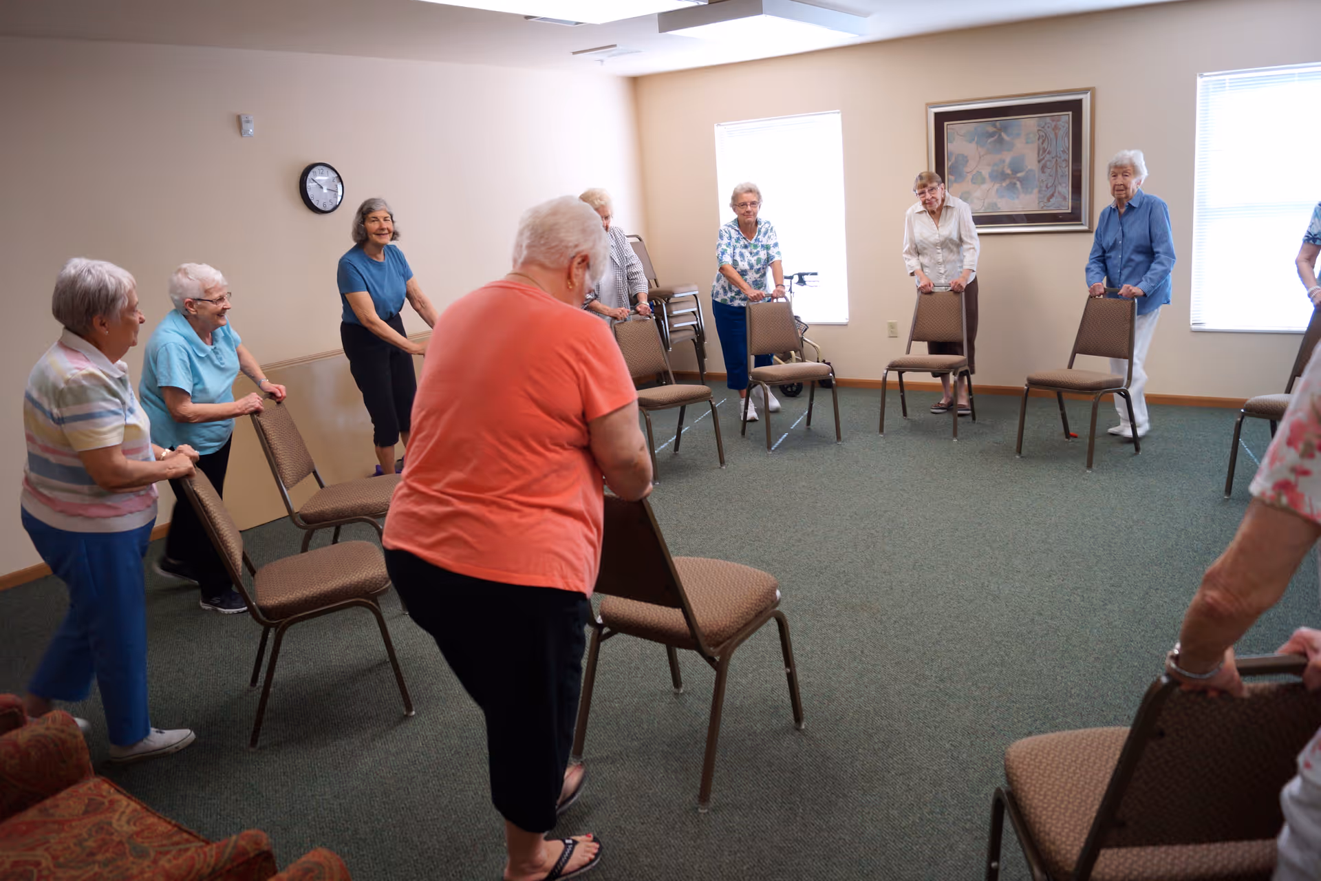 A group of elderly women standing in a circle in a carpeted room, each holding onto the back of a chair, participating in a seated exercise or stretching activity. The room has beige walls, two windows with blinds, a clock on the wall, and a framed picture.