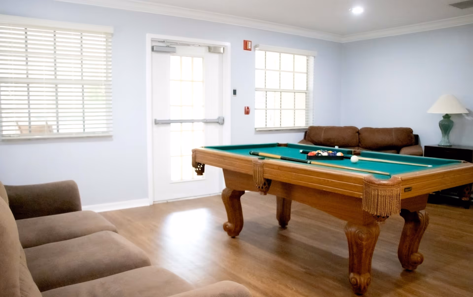 A bright room with a pool table in the center, two brown sofas on opposite sides, a green table lamp on a side table, and windows with white blinds letting in natural light.