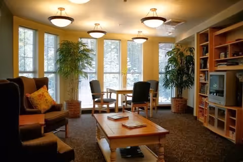 A cozy living room area with large windows letting in natural light. The room features comfortable armchairs with cushions, a wooden coffee table with magazines, a small dining table with four chairs, tall potted plants, and a wooden shelving unit holding a TV and books.