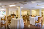 Bright dining room with round tables draped in white linens, wooden chairs, columns, and potted plants.