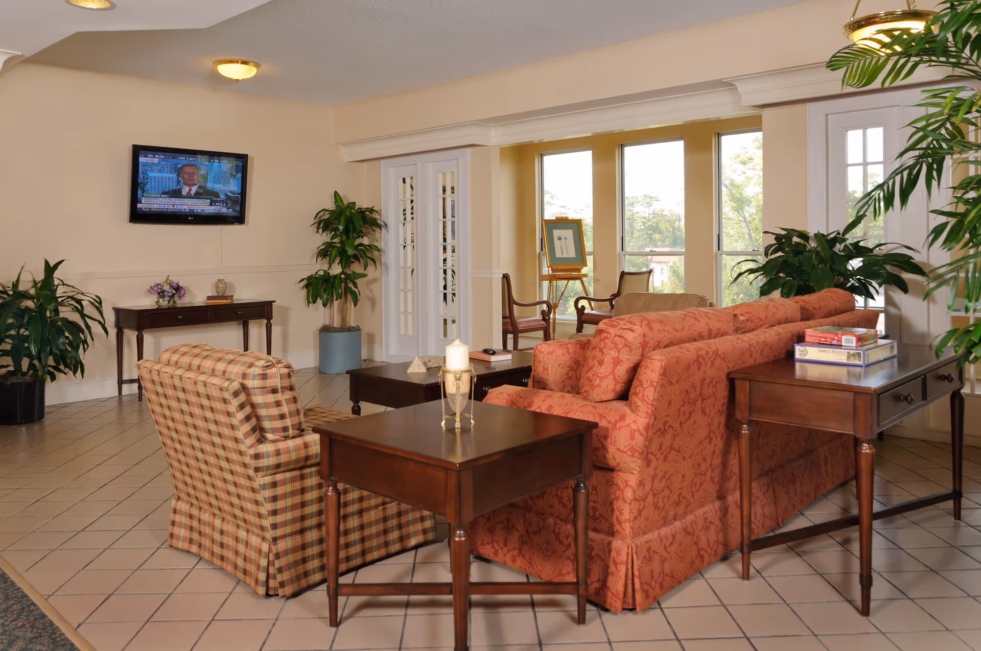 A cozy living room area in a nursing center with a patterned red sofa, a plaid armchair, wooden side tables, and a flat-screen TV mounted on the wall. The room has large windows letting in natural light, several green plants, and a small table with board games on it.