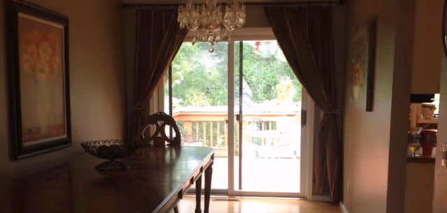 Dining room with a wooden table and chandelier facing a sliding glass door that opens to a deck and trees.