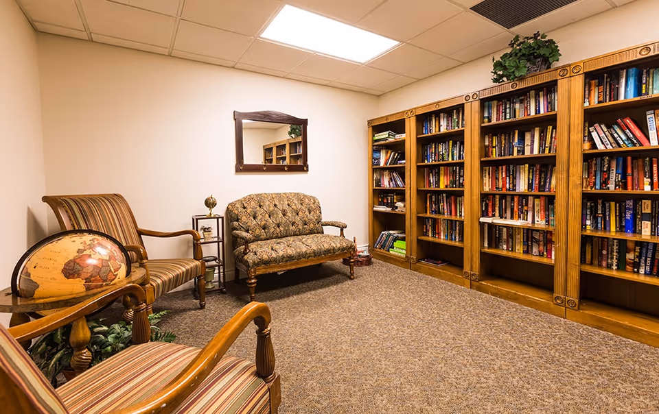 A cozy reading room with a patterned loveseat, two striped armchairs, a wooden bookshelf filled with books, a small side table with a globe, and a mirror on the wall. The room has beige walls and carpeted flooring.
