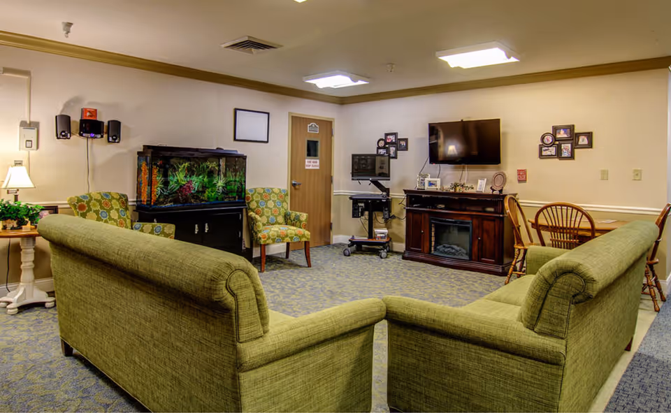 A cozy living room with two green sofas facing a wall-mounted flat screen TV above a wooden electric fireplace. There are two patterned armchairs near a large fish tank on a black stand. A small wooden table with a lamp and a plant is on the left side. On the right, there is a wooden table with chairs. The walls have framed pictures and a door with a sign. The carpet is patterned in blue and beige tones.